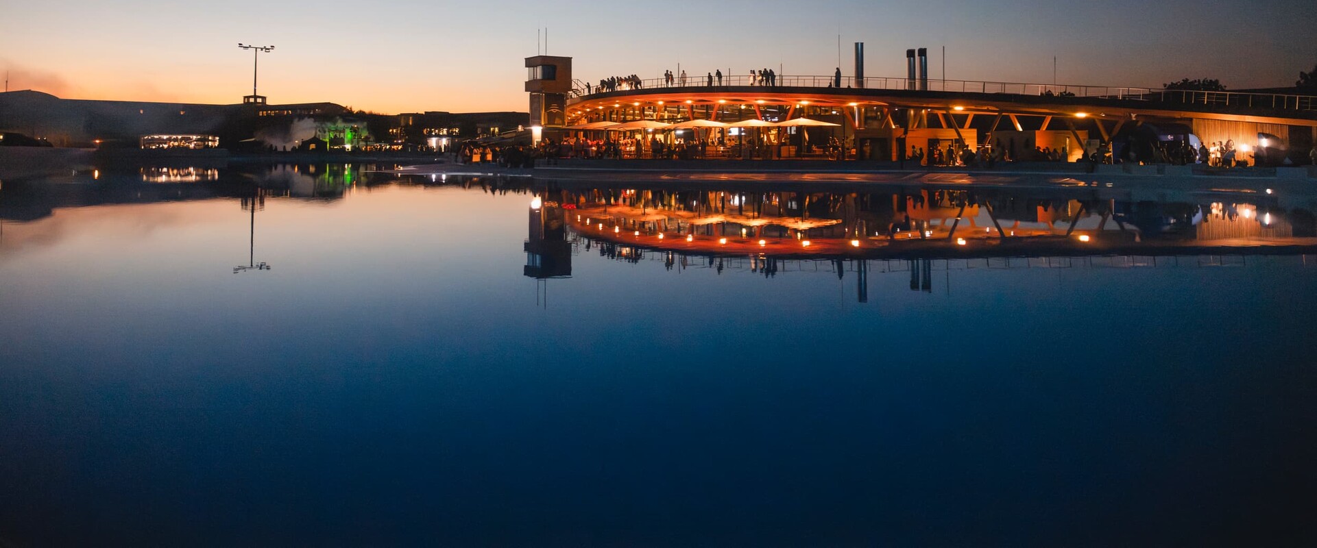 Ob Surfer, Besucher oder Gast im Restaurant oder auf einem Event, die Surftown in M&uuml;nchen bietet Erlebnisse, an die man sich erinnert. Das Bild zeigt eine Aufnahme des Gel&auml;ndes bei D&auml;mmerung, das Wasser der Surfanlage liegt spiegelglatt da und wirft die Reflexion des beleuchteten Geb&auml;udekomplexes mit Restaurant und Walkway &uuml;ber die Rooftop-Terrace. An der Br&uuml;stung sieht man die Silhouetten von Besuchern, die die Aussicht genie&szlig;en.